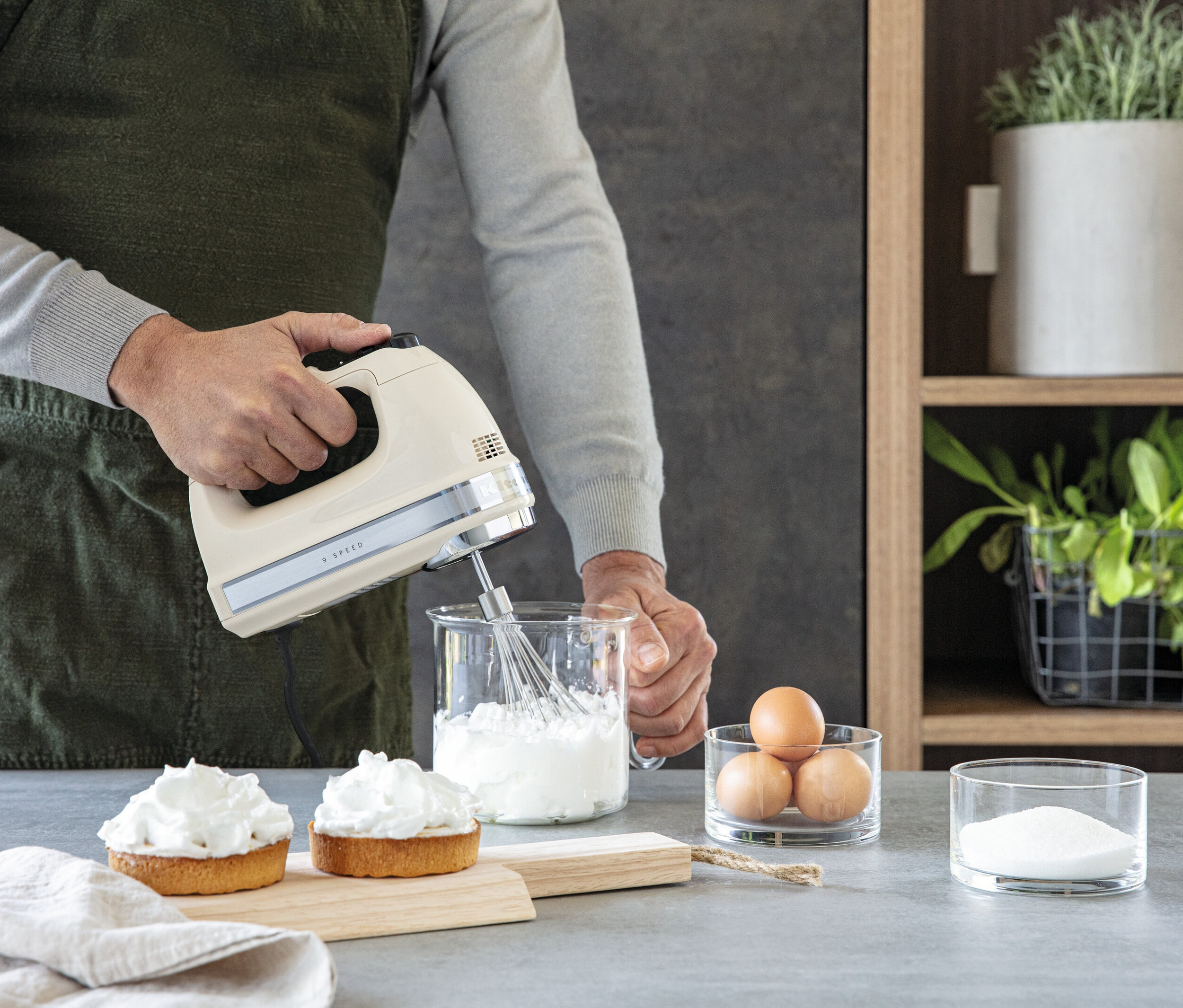 Ein Mann mit Schürze verwendet einen cremefarbenen Handmixer, um Eiweiß in einem Messbecher aus Glas zu schlagen. Auf der Arbeitsfläche befinden sich zwei Törtchen, Eier und Zucker.