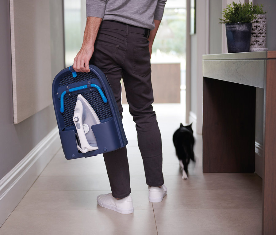Mann trägt ein Bügeleisen in einem blauen Koffer. Hinter ihm geht eine Katze.