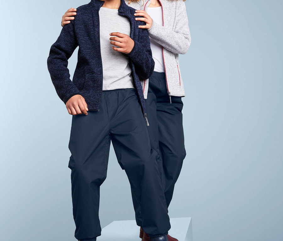 Zwei Kinder stehen auf einer weißen Plattform. Ein Junge mit blauer Jacke und Regenhose. Ein Mädchen mit grauer Jacke und Regenhose.