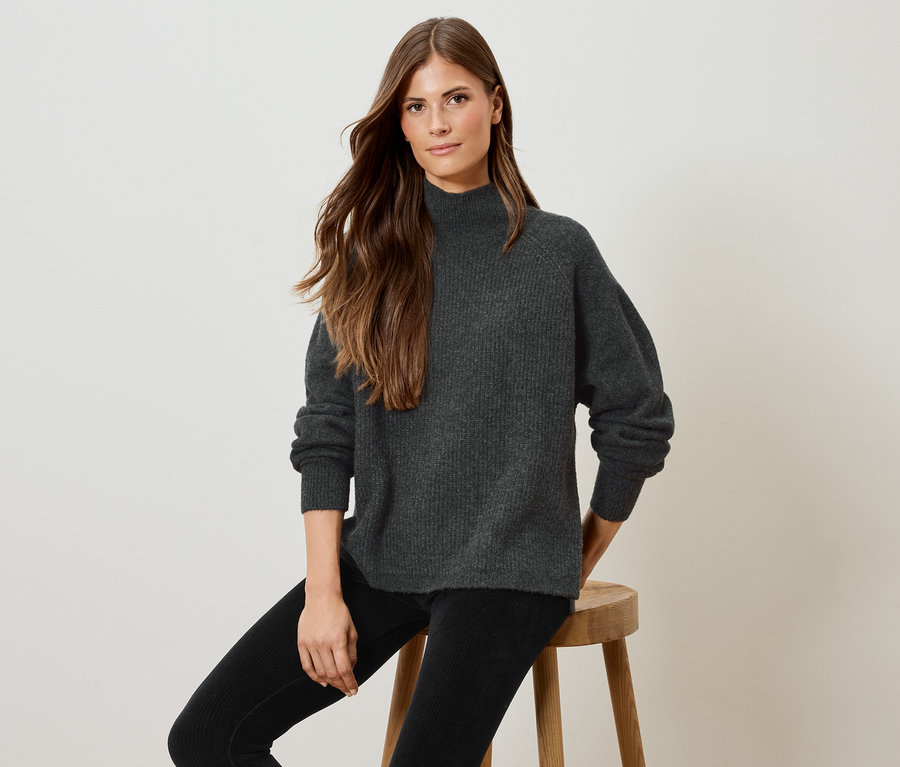 Frau sitzt auf einem Holzhocker und trägt einen grauen Pullover und eine schwarze Hose.