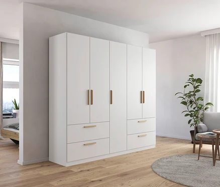 Weißer, fünftüriger Drehtürenschrank Skandi steht im Schlafzimmer neben einem grauen Sessel.