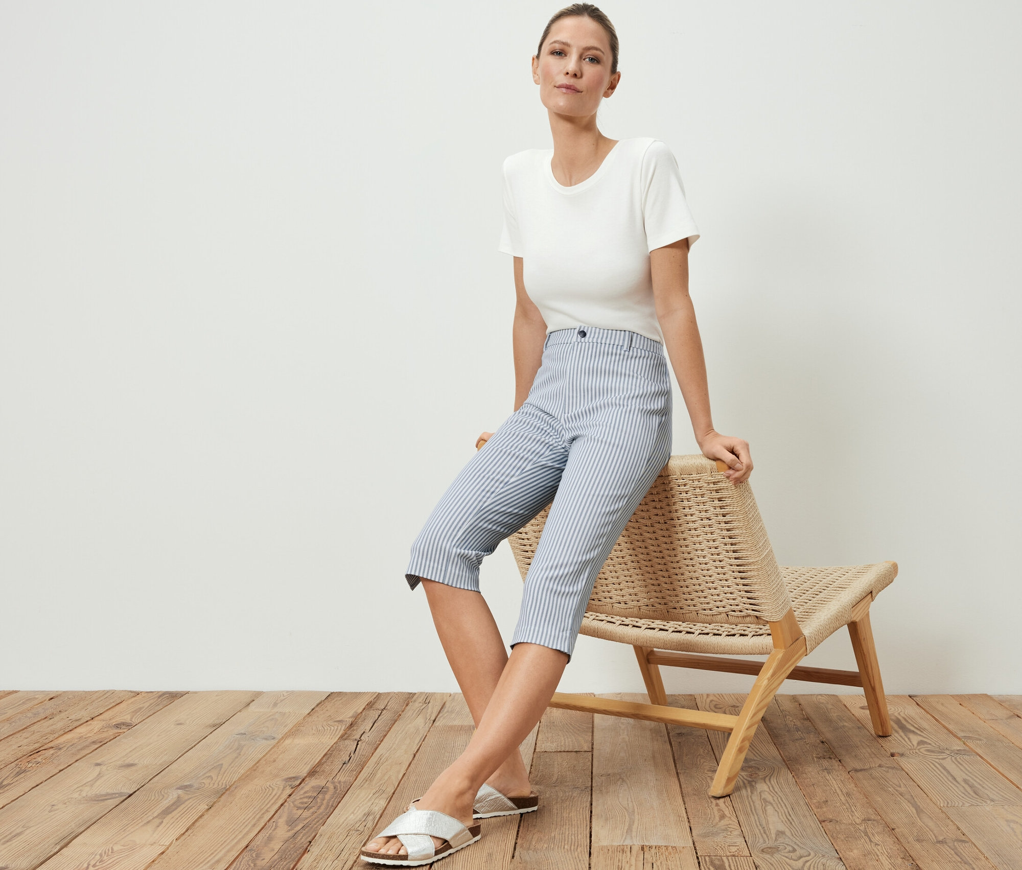 Frau sitzt auf Korbsessel, trägt 3/4-Stretch-Twill-Hose, T-Shirt und Sandalen.