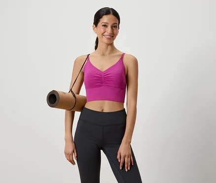 Frau mit brauner Yogamatte über der Schulter, gekleidet in einem pinkfarbenen Sportoberteil und schwarzen Leggings.