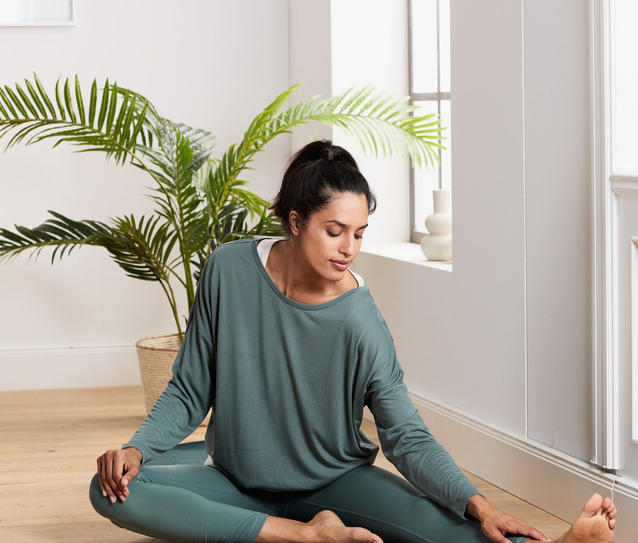 Frau sitzt auf einer Matte und dehnt sich, sie trägt ein grün-weißes 2-in-1-Sportshirt.