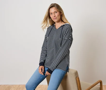 Eine Frau sitzt auf einem Stuhl, sie trägt blaue Jeans und ein gestreiftes Kapuzensweatshirt.
