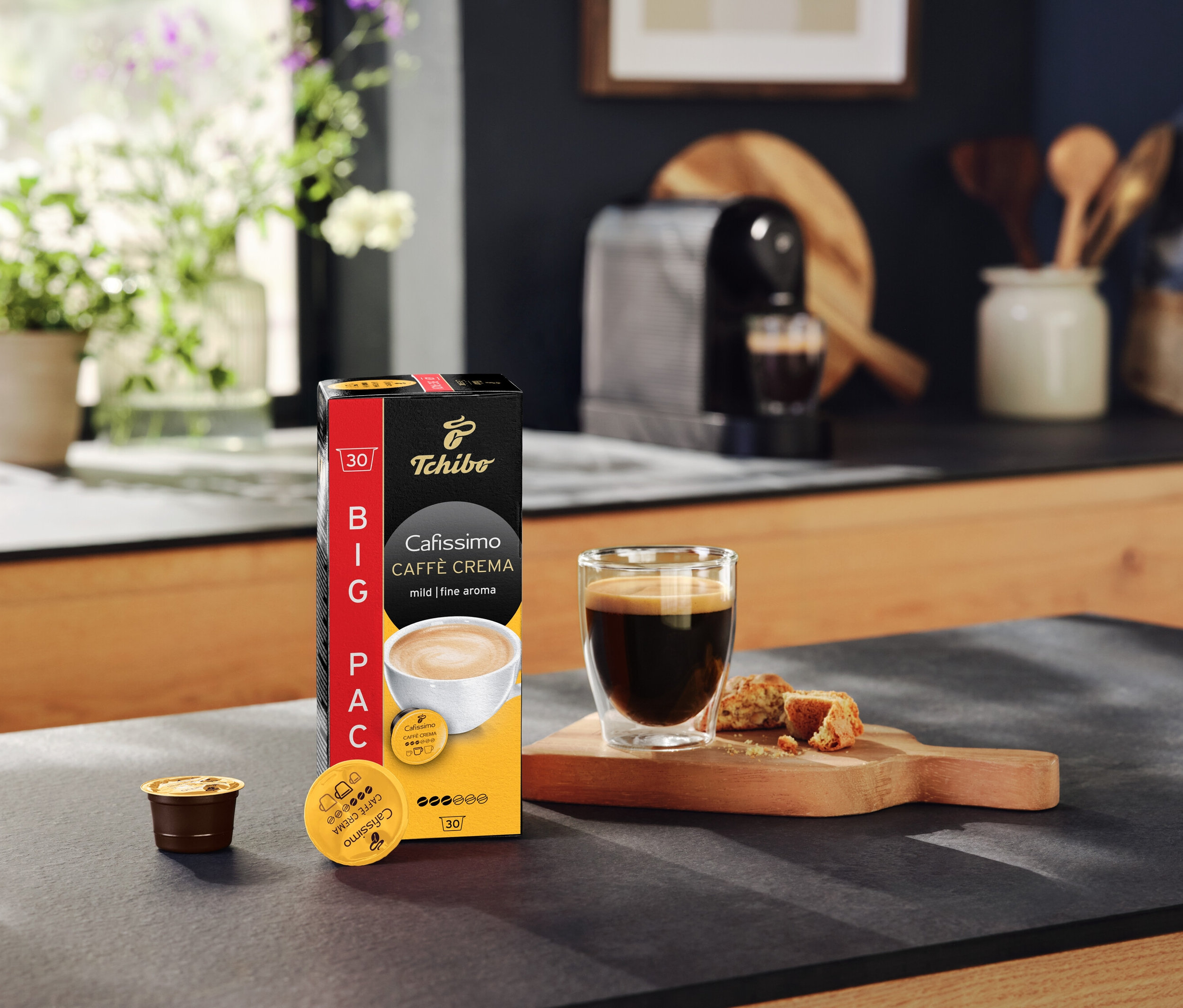 Packung Cafissimo Caffè Crema mild 30 Kapseln, eine Tasse Kaffee und eine Kaffeemaschine auf einer Küchentheke.