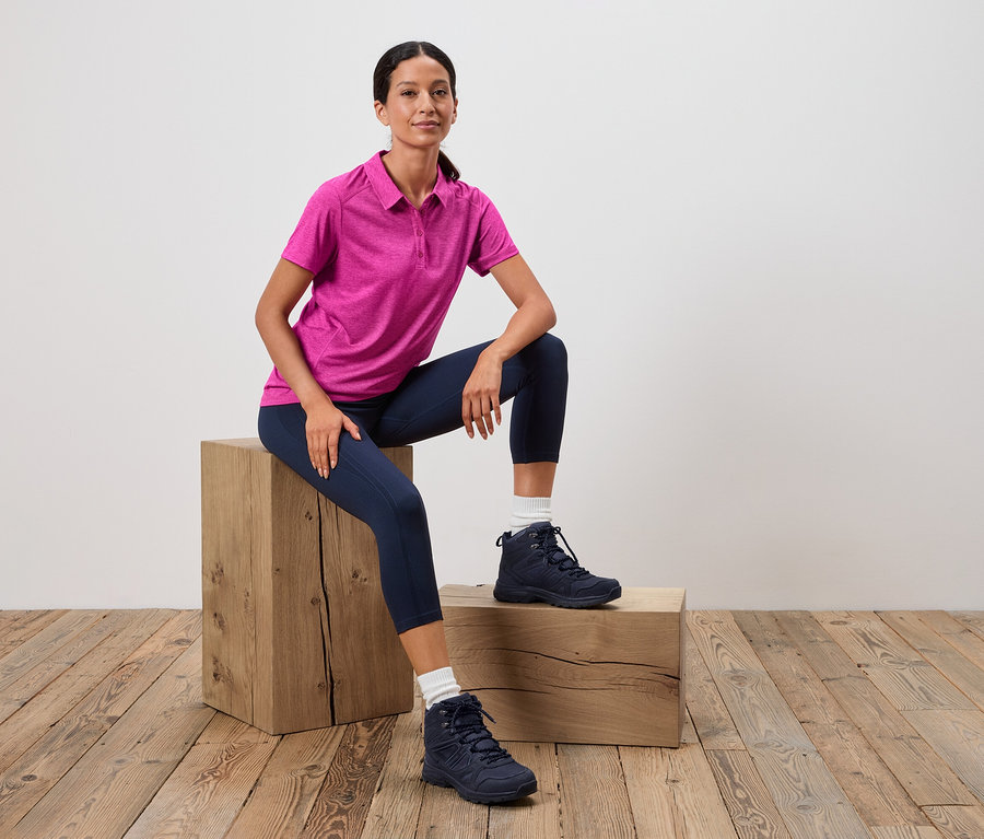 Frau sitzt auf einem Holzschemel, trägt ein pinkes Poloshirt, blaue Leggings und blaue Wanderschuhe.