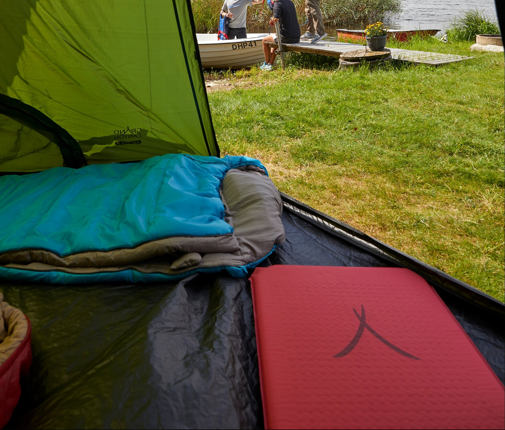 Aus dem Zelt sind drei Personen am See zu sehen. Im Vordergrund befindet sich eine Grand Canyon Isomatte »Hancock 7.5 XW«, ein blauer Schlafsack und eine rote Isomatte.