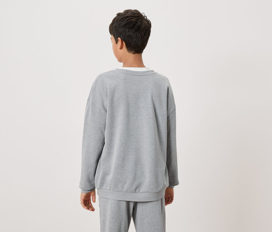 Ein Kind mit dunklen Haaren trägt ein graues Sweatshirt und graue Jogginghose, von hinten gesehen.