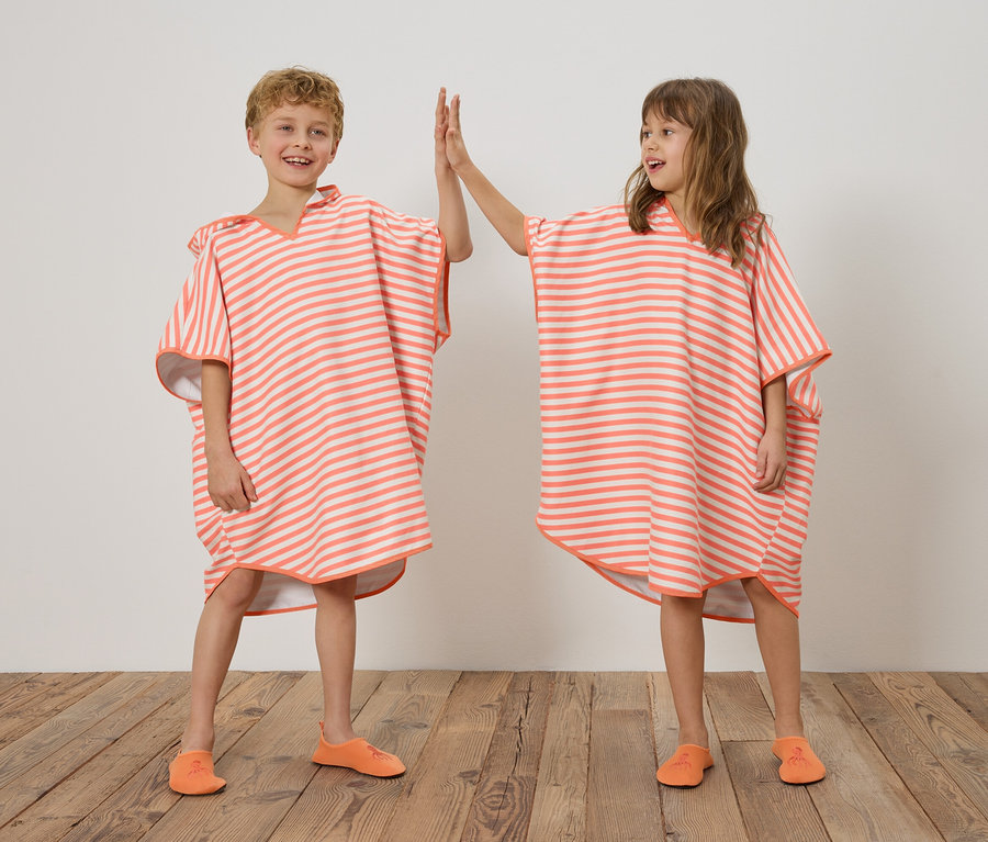 Zwei Kinder, ein Junge und ein Mädchen, stehen auf einem Holzboden vor einer weißen Wand und geben sich ein High-Five. Beide tragen gestreifte Ponchos in Orange und Weiß sowie orangefarbene Schuhe.