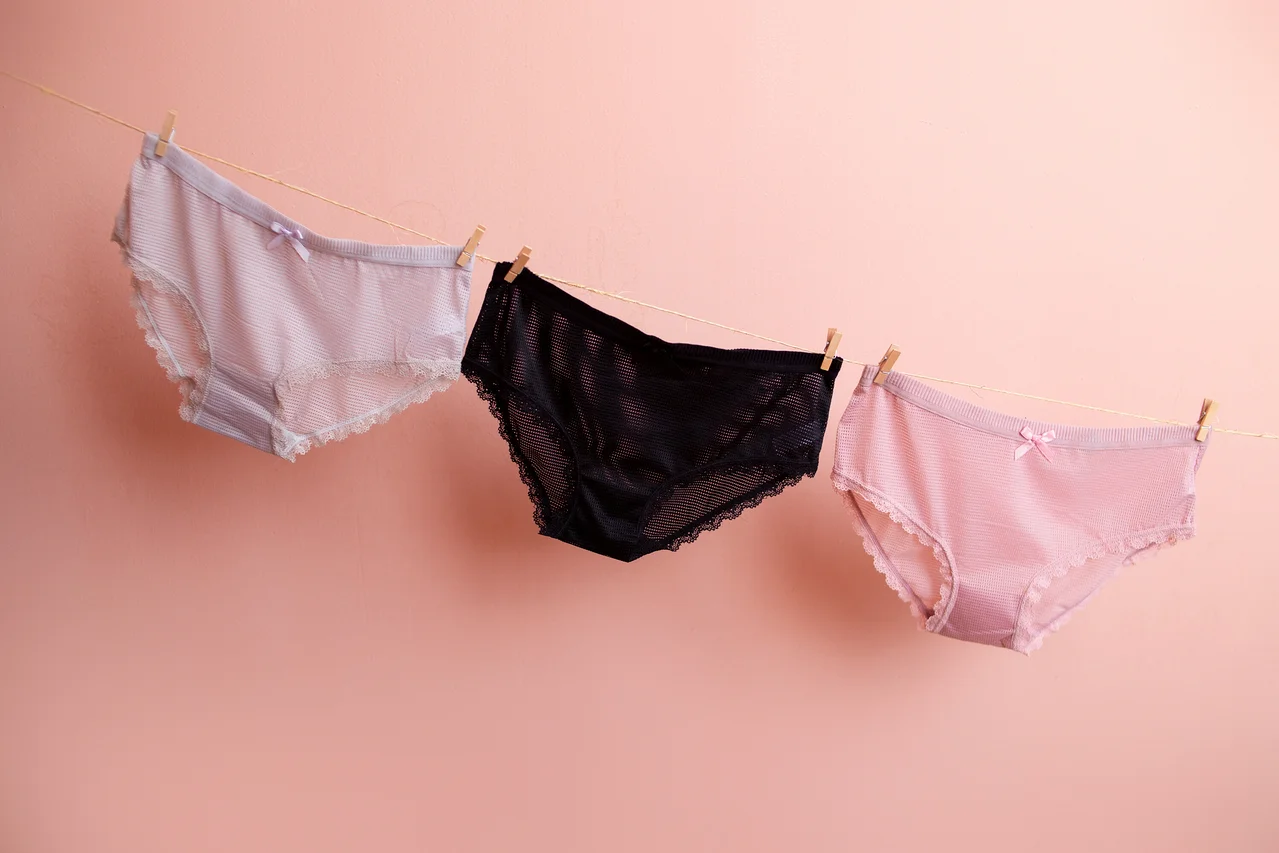 Drei Damenunterhosen in Rosa, Weiß und Schwarz hängen an einer Wäscheleine vor einem rosa Hintergrund.