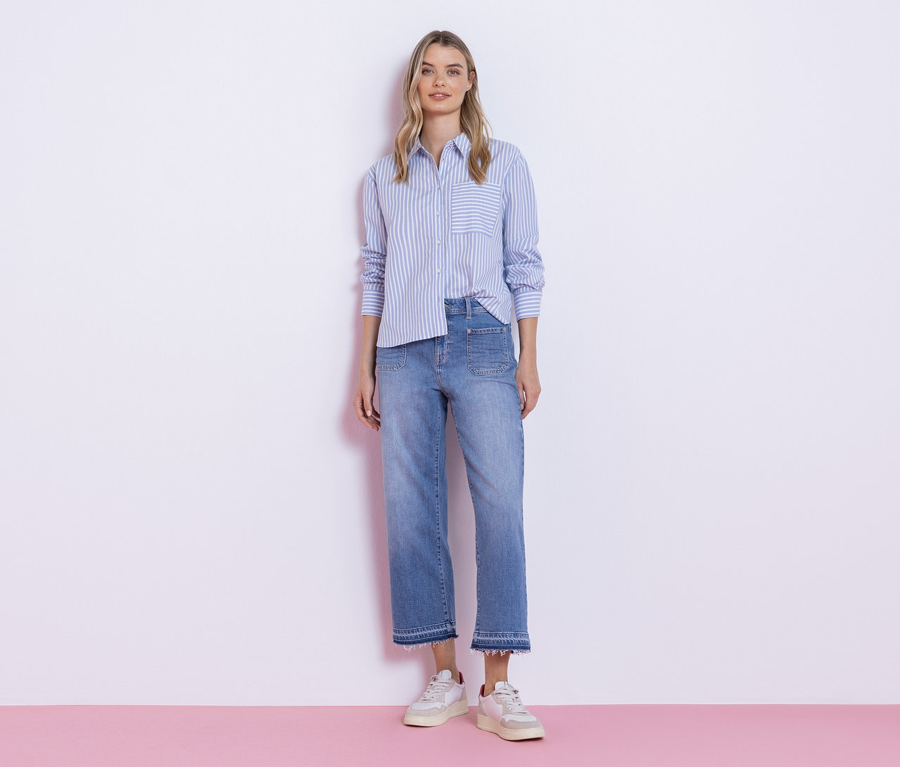 Eine junge Frau steht vor einer weißen Wand und trägt eine blau-weiß gestreifte Street One Hemdbluse, eine helle Jeans mit ausgefranstem Saum und weiße Sneaker.