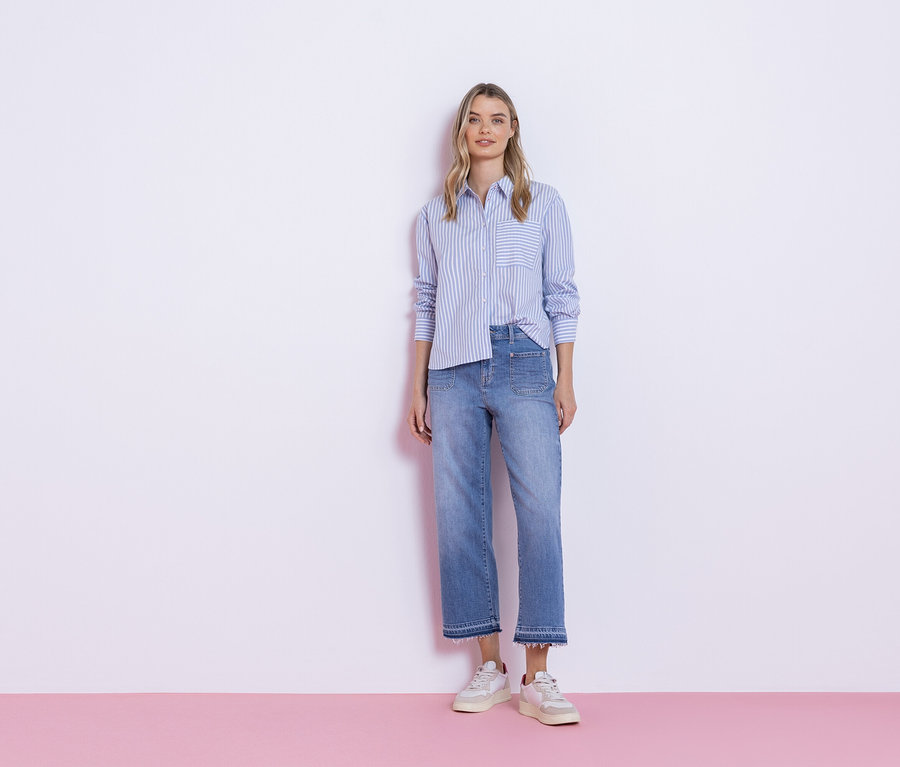 Eine Frau steht vor einer weißen Wand, gekleidet in ein blau gestreiftes Hemd, Jeans und Turnschuhe.