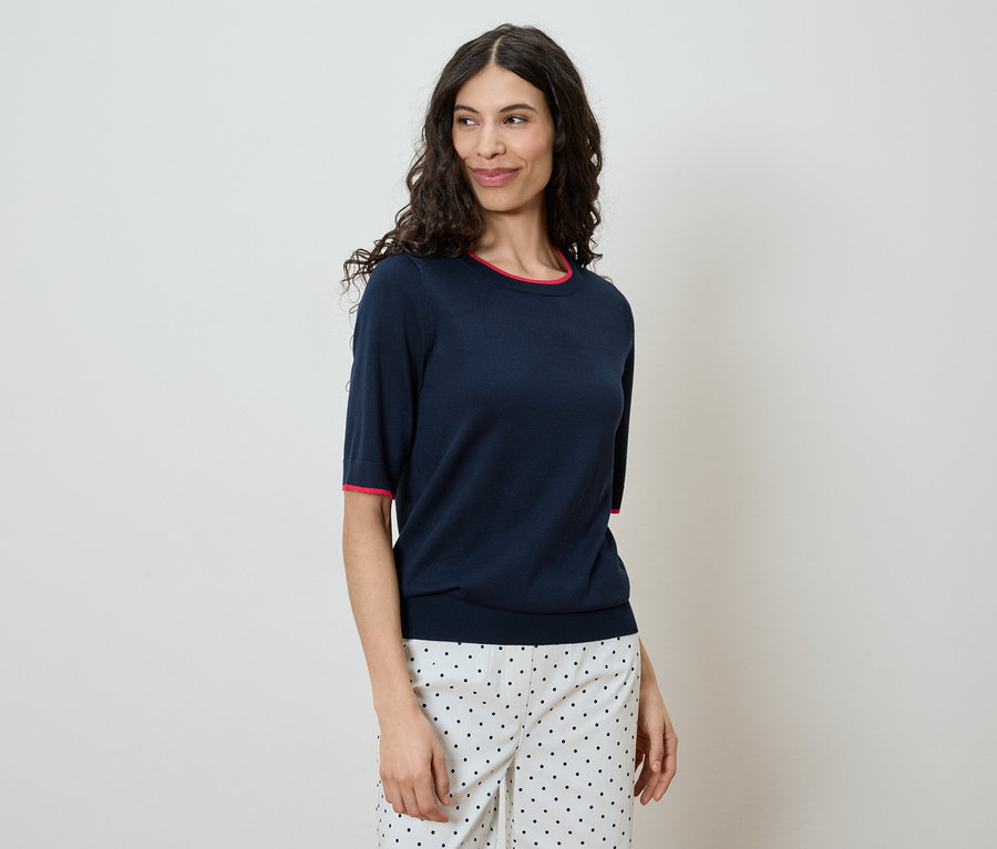 Frau mit gewelltem, dunklem Haar trägt marineblaues Kurzarm-Shirt mit rotem Saum und gepunktete Hose vor einem hellen Hintergrund.