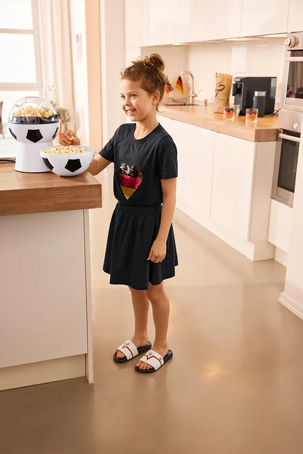 Mädchen isst Popcorn neben Fußball-Popcornmaschine. Sie trägt Kinder-T-Shirt mit Wendepailletten, Herz, Kinder-Jerseyrock mit Hose und Badelatschen.