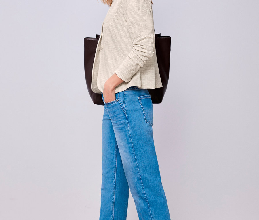 Frau trägt einen cremefarbenen Street One Jerseyblazer, blaue Jeans und dunkle flache Schuhe, eine Hand in der Tasche, mit einer dunklen Tragetasche.