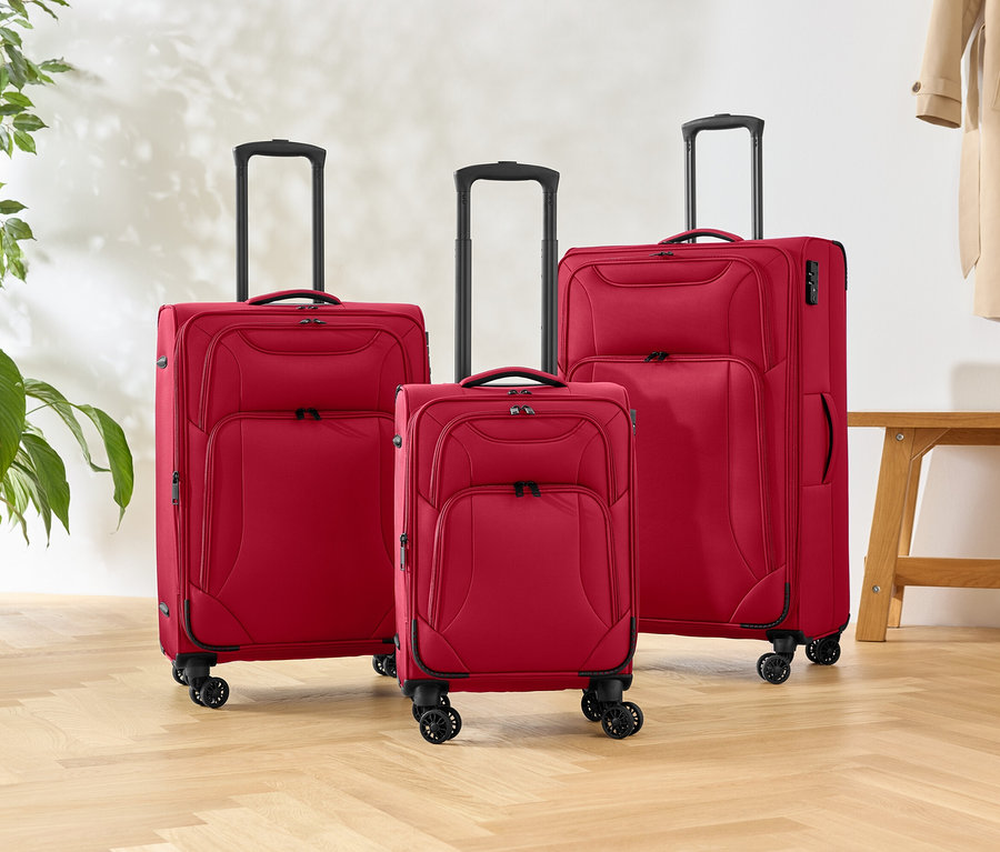 Drei rote Rollkoffer unterschiedlicher Größe stehen auf einem Holzboden, daneben eine Grünpflanze und eine Bank.