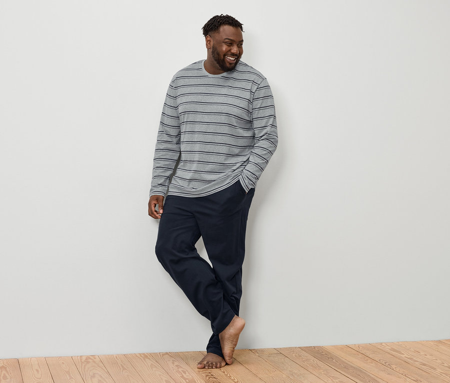 Ein lächelnder Mann mit dunklen Haaren und Bart steht barfuß da, bekleidet mit einem grau gestreiften Langarmshirt und marineblauen Hosen.