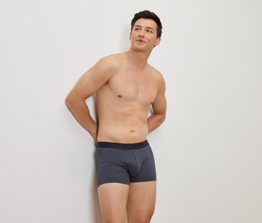 Mann mit freiem Oberkörper lehnt sich an eine weiße Wand und trägt blaue Boxershorts mit Muster.
