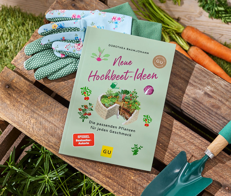 Gartenhandschuhe, Karotten, eine grüne Gartenschaufel und das Buch 'Neue Hochbeet-Ideen' liegen auf einer Holzkiste.
