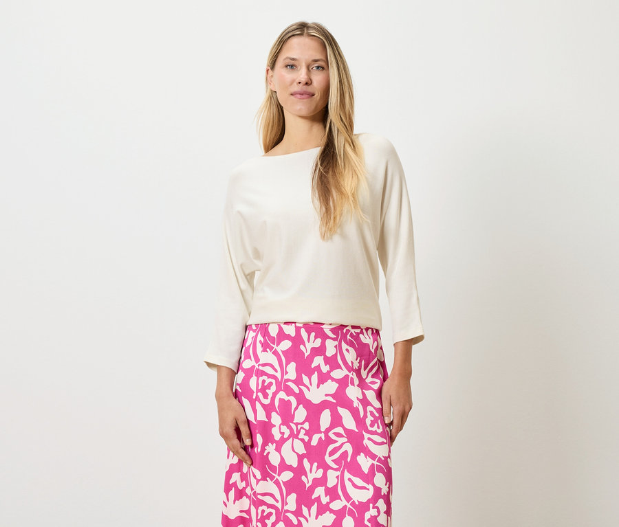 Eine Frau mit blonden Haaren trägt ein cremefarbenes Oberteil und einen pinkfarbenen Rock mit Blumendruck.
