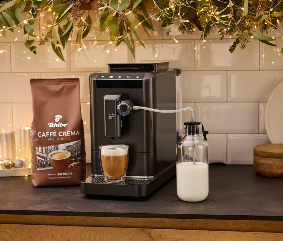 Ein »Esperto 2 Milk« Kaffeevollautomat, Granite Black mit einer Tasse Kaffee und einer Packung Kaffee Crema. Milch wird in einem Glasbehälter aufbewahrt.