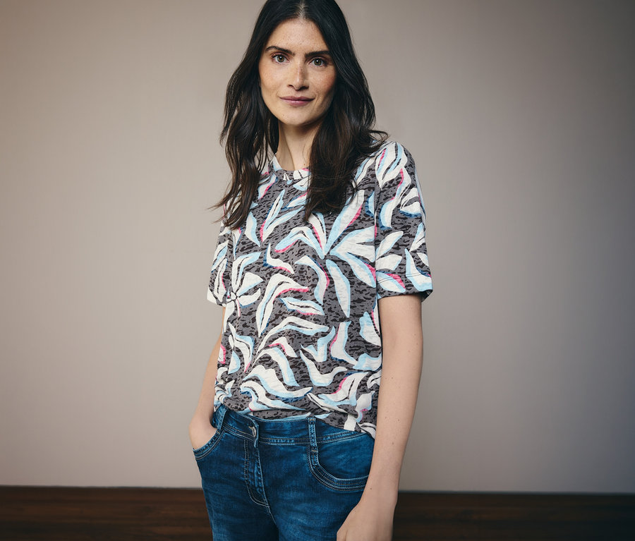 Frau mit dunklen Haaren trägt ein blaues CECIL Kurzarmshirt mit Muster und blaue Jeans.