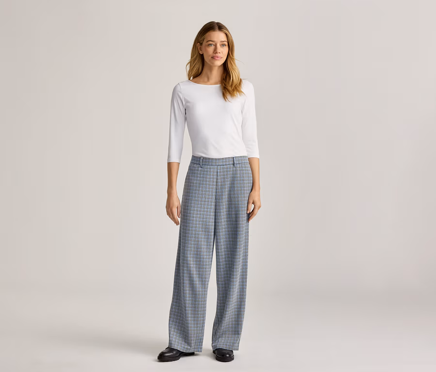 Eine Frau steht in einem weißen, langärmeligen Oberteil und einer weiten, blau karierten Hose mit schwarzen Schuhen.