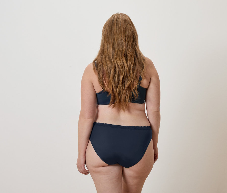 Rückansicht einer Frau mit langen, gewellten, rötlich-blonden Haaren, die einen marineblauen BH und passende Unterwäsche trägt.