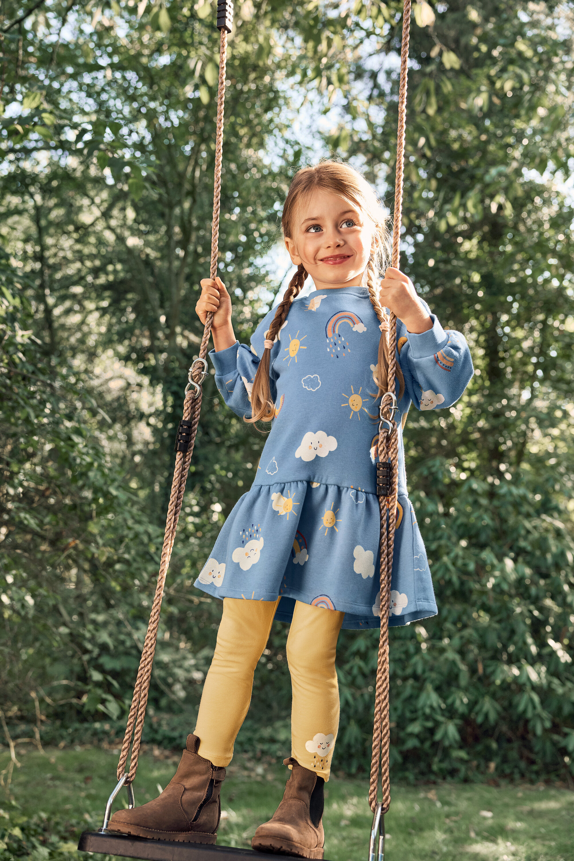Ein junges Mädchen mit Zöpfen schwingt auf einer Schaukel und trägt ein blaues Kleid mit einem Muster aus Sonnen, Wolken und Regenbögen, gelbe Leggings und braune Stiefel.