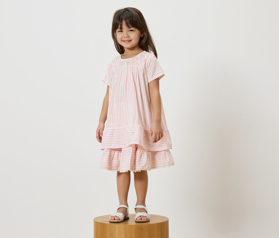 Mädchen steht auf einem Hocker und trägt ein rosa gestreiftes Kleid mit weißen Sandalen.