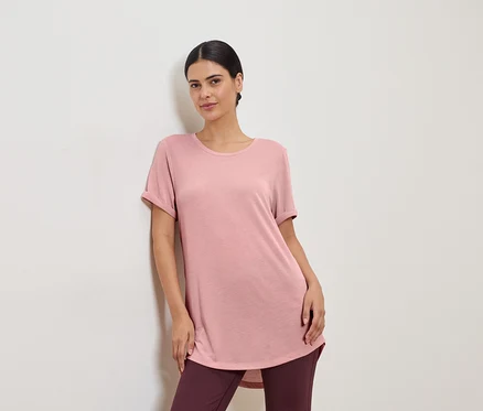 Frau in rosafarbenem T-Shirt und Leggings, die an einer weißen Wand lehnt.