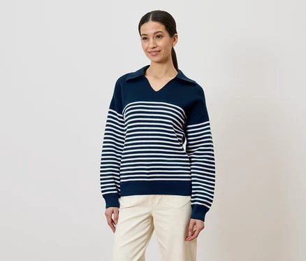 Frau mit Pferdeschwanz trägt einen dunkelblauen, gestreiften Pullover und eine helle Hose vor einem weißen Hintergrund.