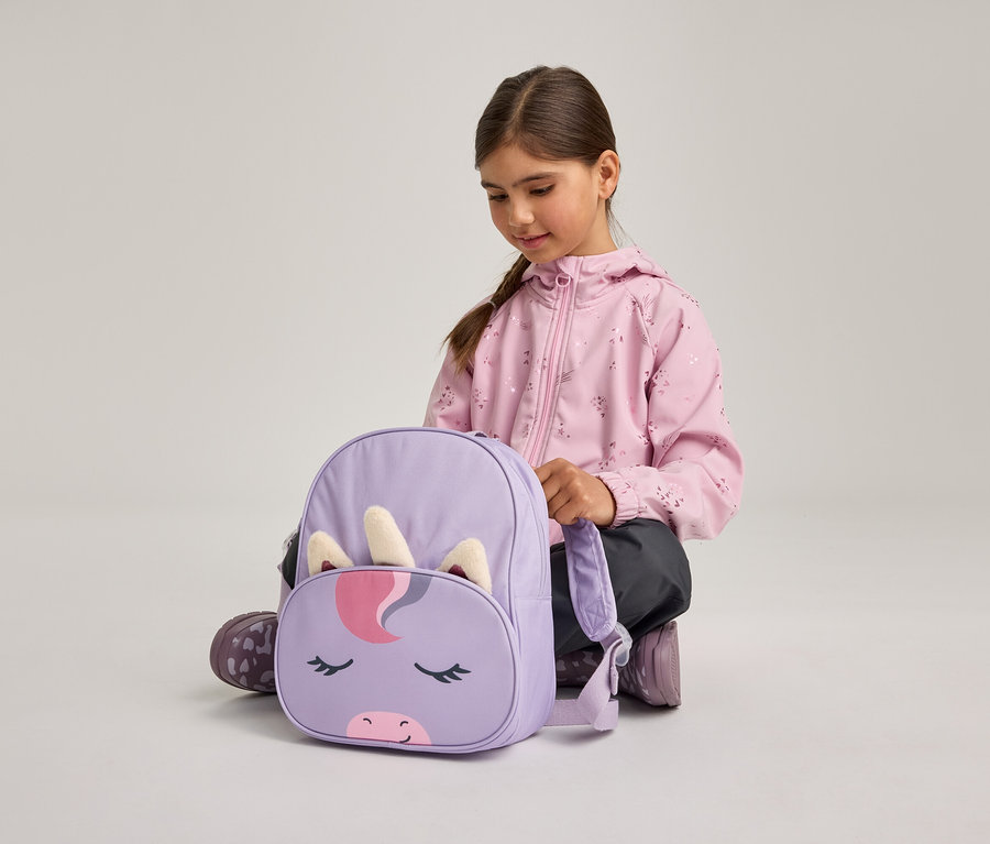 Ein junges Mädchen in einer rosa Jacke und dunklen Hosen hockt am Boden und blickt auf einen lila Rucksack mit Einhorn-Gesichtsdesign, inklusive Horn und Ohren.