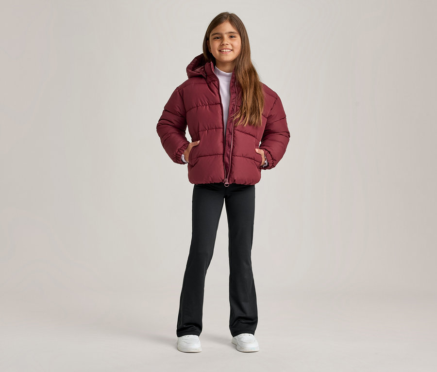 Lächelndes Mädchen in bordeauxroter Steppjacke mit Kapuze, schwarzer Schlaghose und weißen Turnschuhen, die Hände in den Jackentaschen.