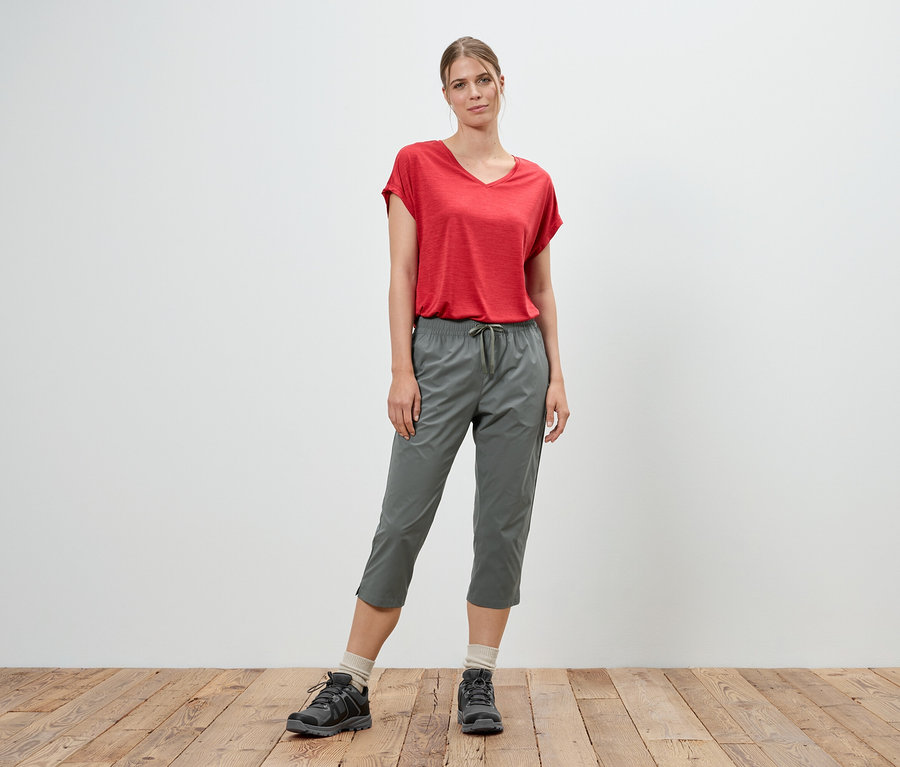 Frau steht in rotem T-Shirt und grauen Dreiviertelhosen vor einer weißen Wand.