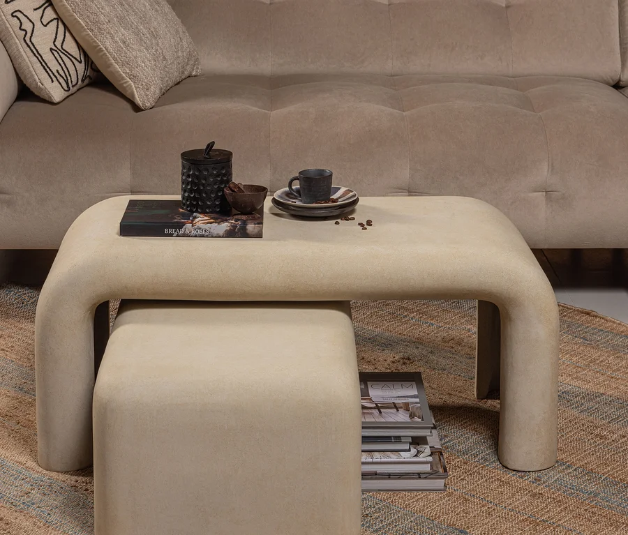Ein beiges, getuftetes Sofa mit Kissen hinter einem hellbeigen, ineinandersteckbaren Couchtisch-Set auf einem gewebten Teppich. Auf dem oberen Tisch liegen ein Buch, ein Deckelgefäß, eine Schale und eine Kaffeetasse; Zeitschriften sind auf dem unteren Tisch gestapelt.