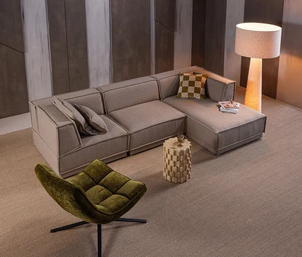 Ein modernes Wohnzimmer mit einem beigen L-förmigen Sofa, einem grünen Sessel, einem strukturierten Beistelltisch mit Glas und Buch sowie einer hohen Stehlampe auf einem beigen Teppich.