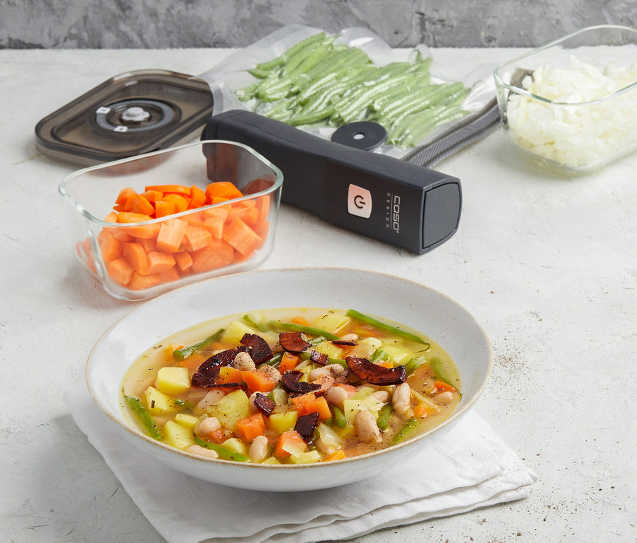 Eine Schüssel Gemüsesuppe mit Kartoffeln, Karotten, grünen Bohnen und weißen Bohnen steht auf einer weißen Serviette. Dahinter liegen ein schwarzer CASO Vacu OneTouch Handvakuumierer, ein Vakuumbeutel mit grünen Bohnen, ein Glasbehälter mit gewürfelten Karotten und einer mit gewürfelten Zwiebeln.