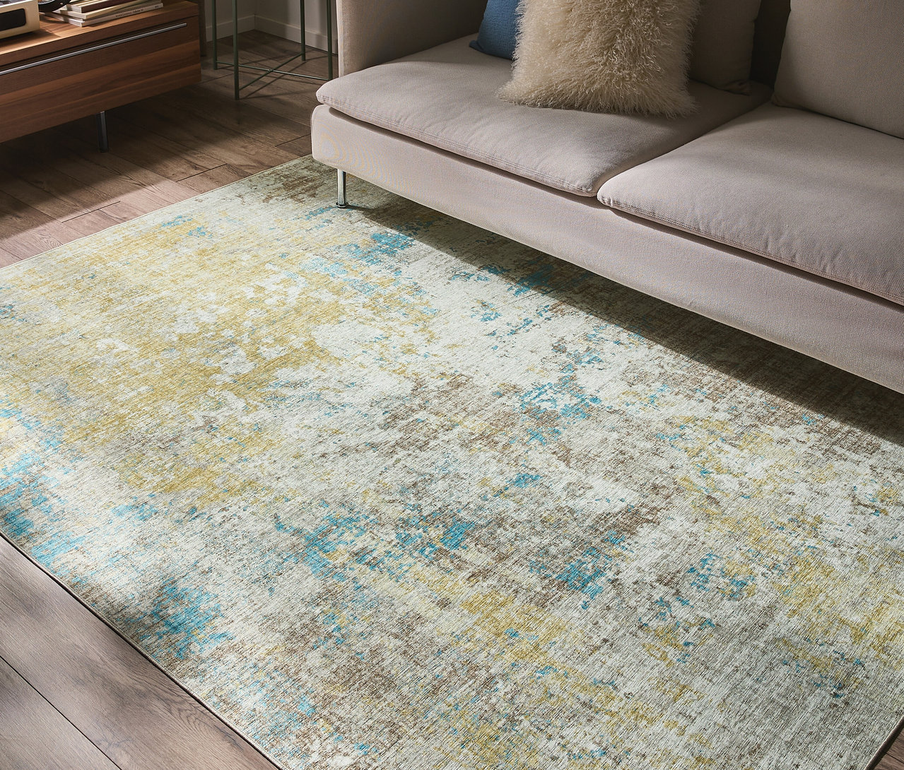 Ein benuta Teppich »Laury« mit abstraktem Muster in Blau, Gelb, Braun und Weiß liegt auf einem Holzboden vor einem hellen Sofa.