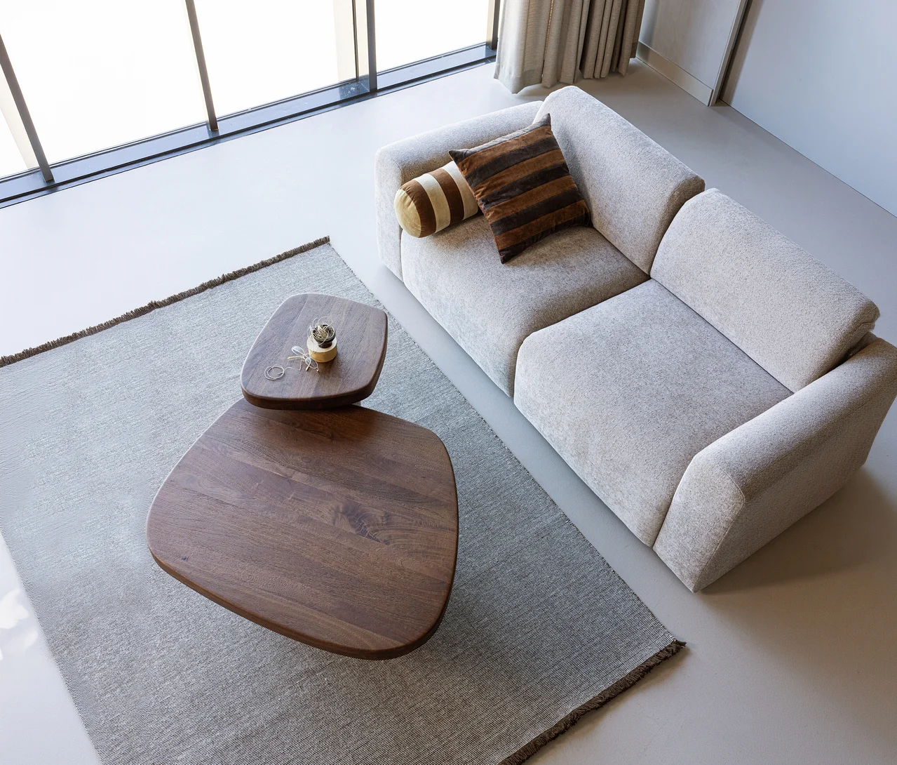 Eine Draufsicht auf ein modernes Wohnzimmer mit einem hellgrauen Sofa mit gestreiften Kissen, zwei organisch geformten Couchtischen aus dunklem Holz und einem braunen WOOOD Teppich »Weavly« auf hellgrauem Boden. Der kleinere Tisch trägt eine kleine Pflanze und einen Ring.