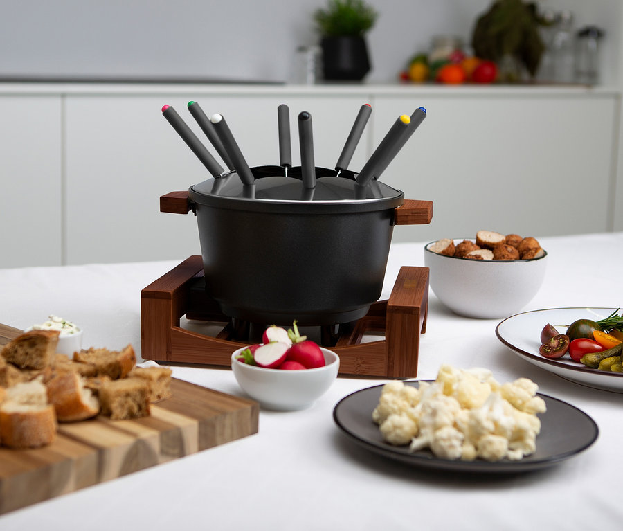 Schwarzes Fondue-Set mit Gabeln auf Holzständer, umgeben von Schüsseln mit Essen.