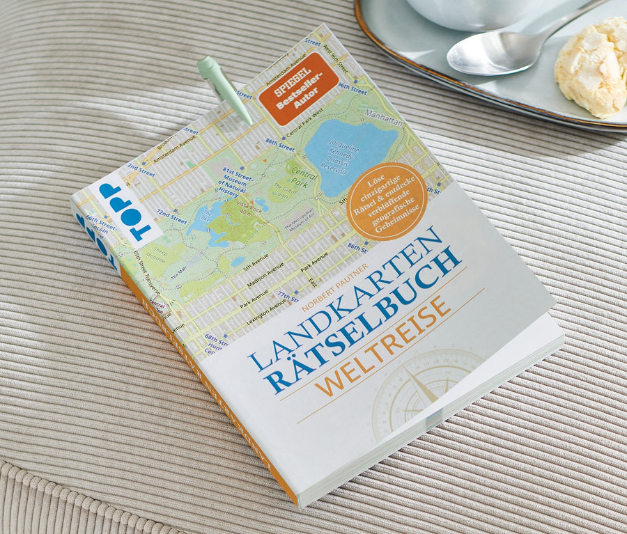 Auf einem beigen Hocker liegt ein Buch: Landkarten-Rätselbuch »Weltreise«, daneben eine Tasse Kaffee und Kekse.