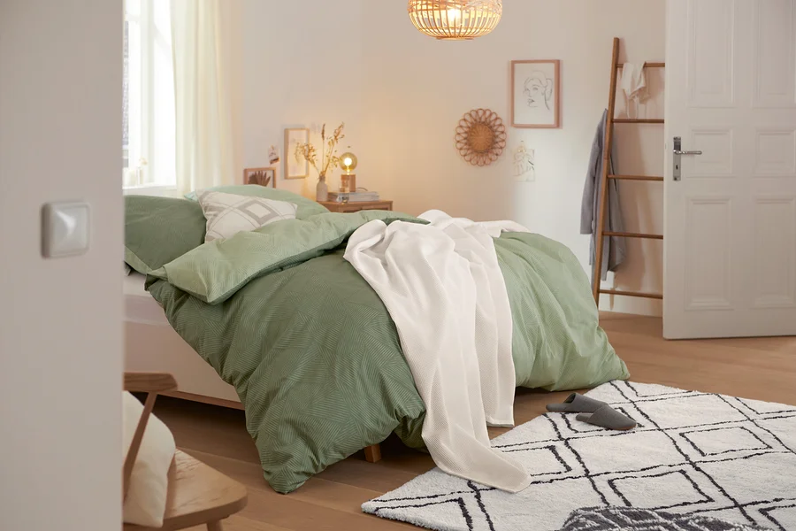 Grüne Renforcé-Bettwäsche und Tagesdecke liegen auf einem Bett. Dekokissen und ein Hochflorteppich sind auch sichtbar.