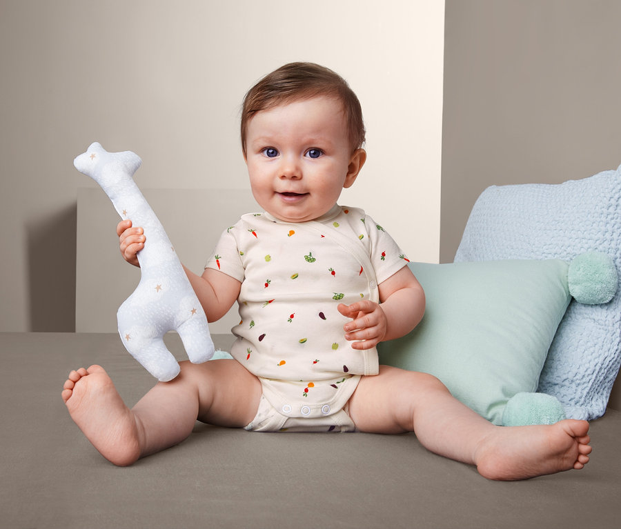 Ein Baby sitzt und hält ein Stofftier-Giraffe. Es trägt einen 3 Kurzarmbody mit Gemüseaufdruck.