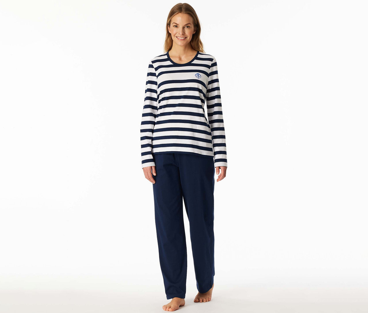 Lächelnde Frau in einem SCHIESSER Pyjama-Set: weiß-marineblau gestreiftes Langarmshirt mit Anker-Logo und marineblaue Hose, barfuß.