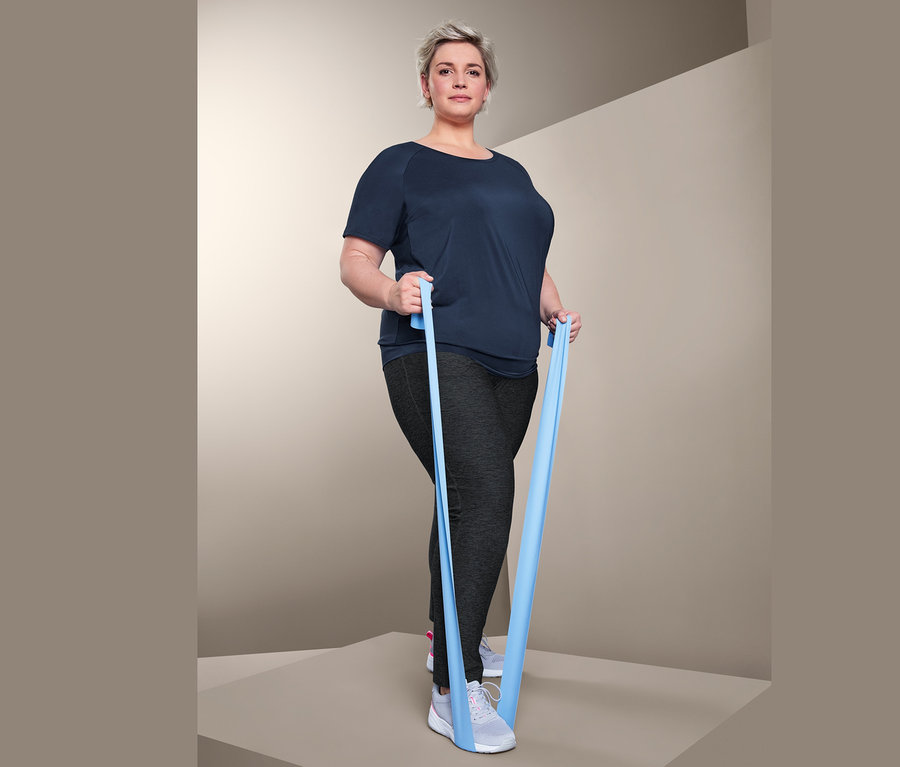 Frau trainiert mit einem blauen Resistance Band. Sie trägt ein Sportshirt und eine Trainingshose.
