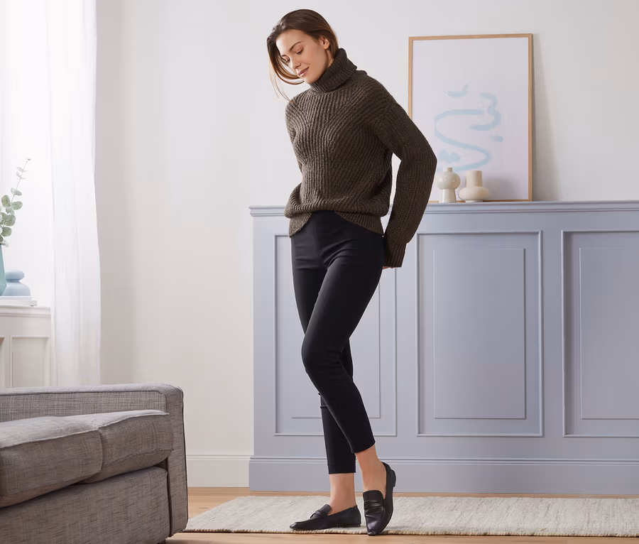 Frau steht im Wohnzimmer und trägt einen braunen Pullover und eine schwarze Stretchhose.