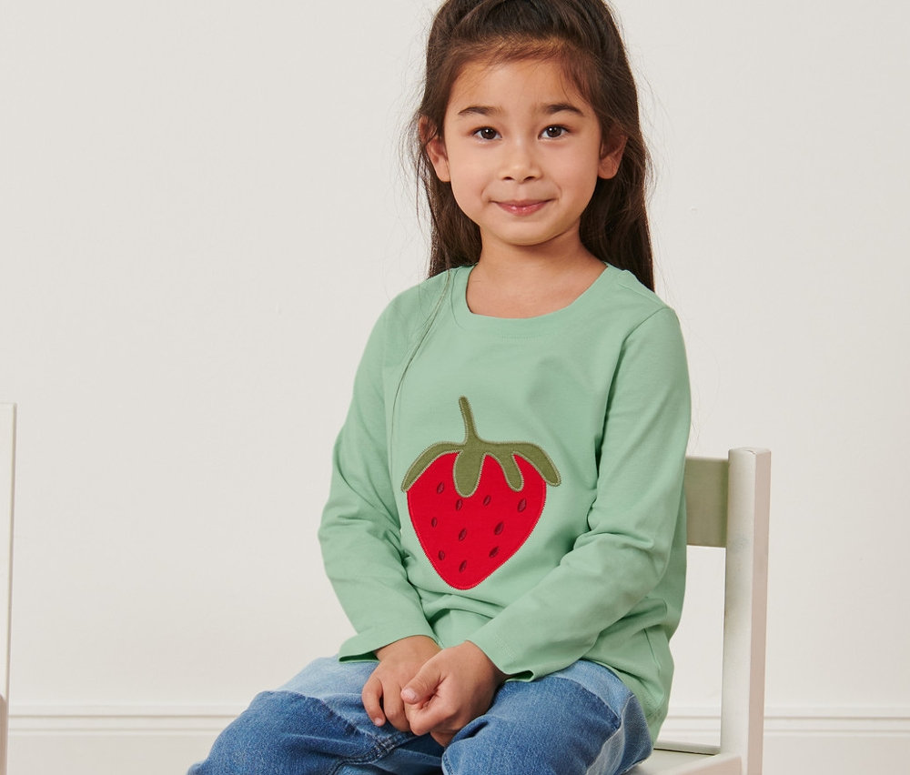 Ein Mädchen sitzt auf einem Stuhl und trägt ein grünes Shirt mit Erdbeermotiv. Im Hintergrund ist eine weiße Wand.