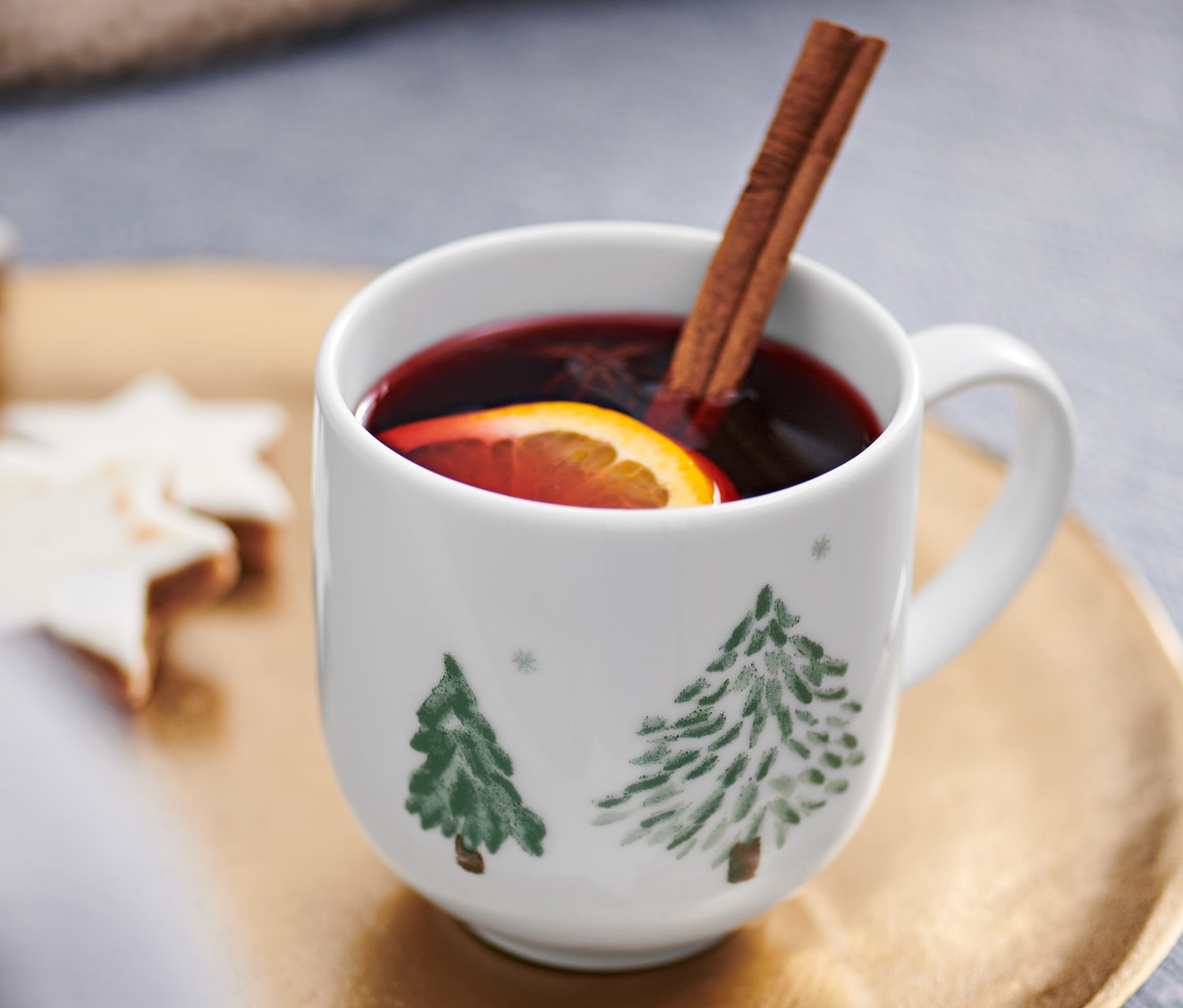 Becher mit heißem Getränk, Orangenscheibe und Zimtstange auf goldenem Tablett neben sternförmigen Keksen.